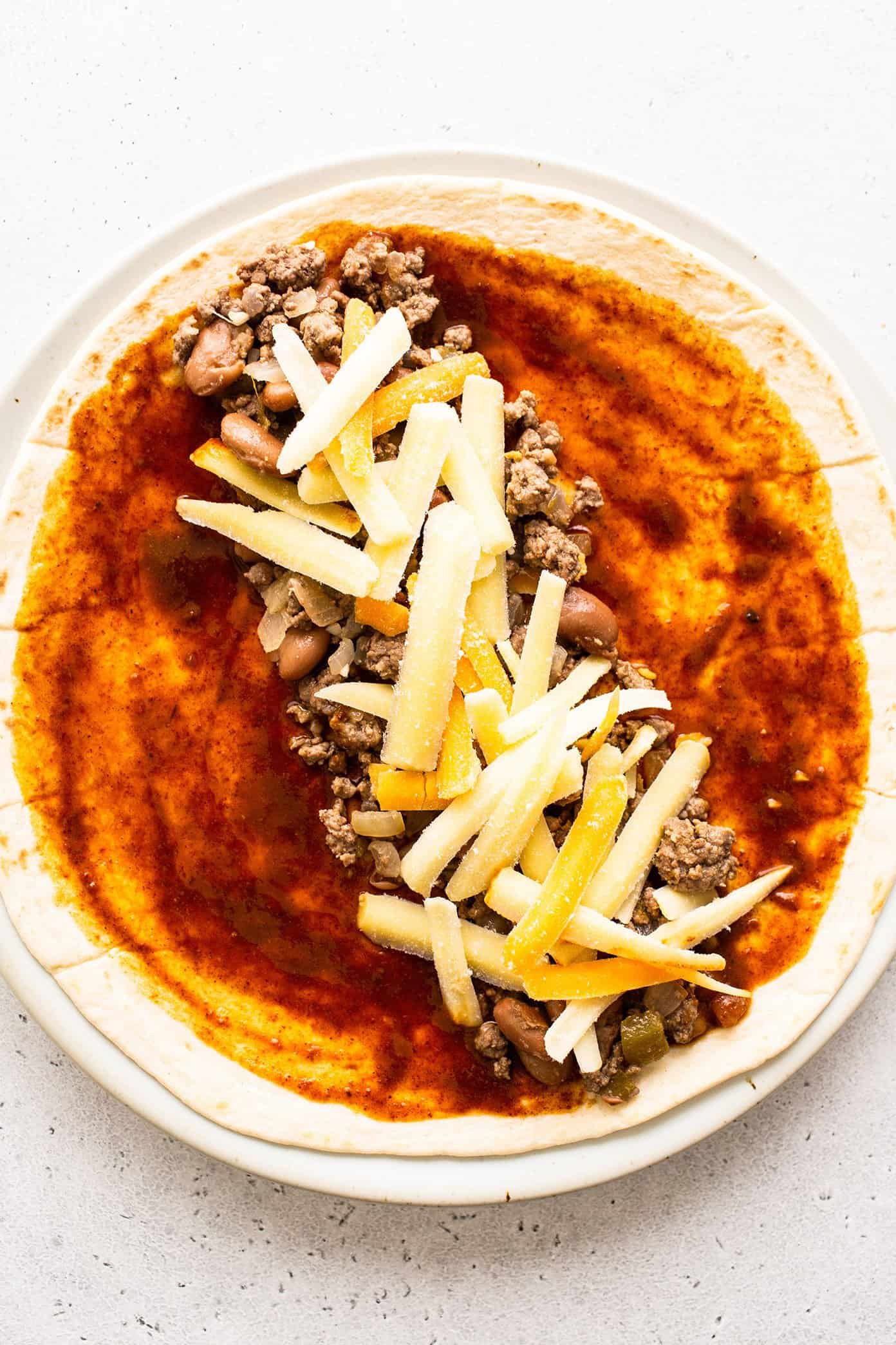 These delicious Tex-Mex-inspired enchiladas are loaded up with with tender ground beef (or plant-based meat), green chiles, onion, garlic, beans, cheese, corn or flour tortillas, and my favorite red enchilada sauce. When baked to warm and melty perfection, and served up with a few slices of avocado and perhaps a sprinkling of fresh cilantro and crumbled cotija cheese, they are always a crowd fave! Freezer and make ahead instructions included. | gimmesomeoven.com Beef enchilada filling spread on a tortilla with cheese and red enchilada sauce