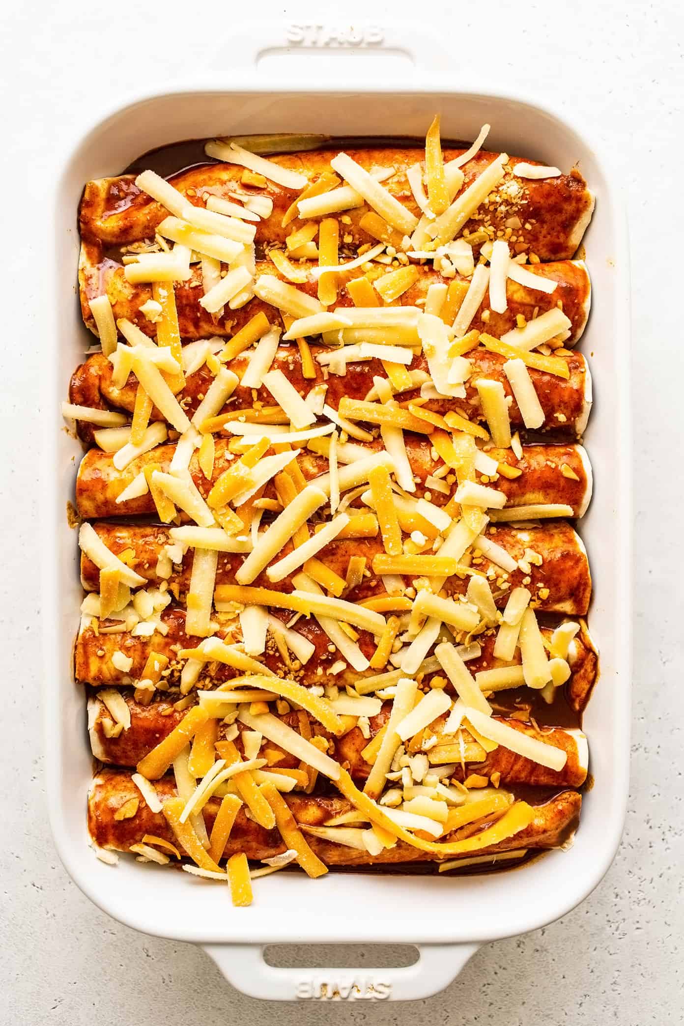 These delicious Tex-Mex-inspired enchiladas are loaded up with with tender ground beef (or plant-based meat), green chiles, onion, garlic, beans, cheese, corn or flour tortillas, and my favorite red enchilada sauce. When baked to warm and melty perfection, and served up with a few slices of avocado and perhaps a sprinkling of fresh cilantro and crumbled cotija cheese, they are always a crowd fave! Freezer and make ahead instructions included. | gimmesomeoven.com Uncooked beef enchiladas in pan with cheese