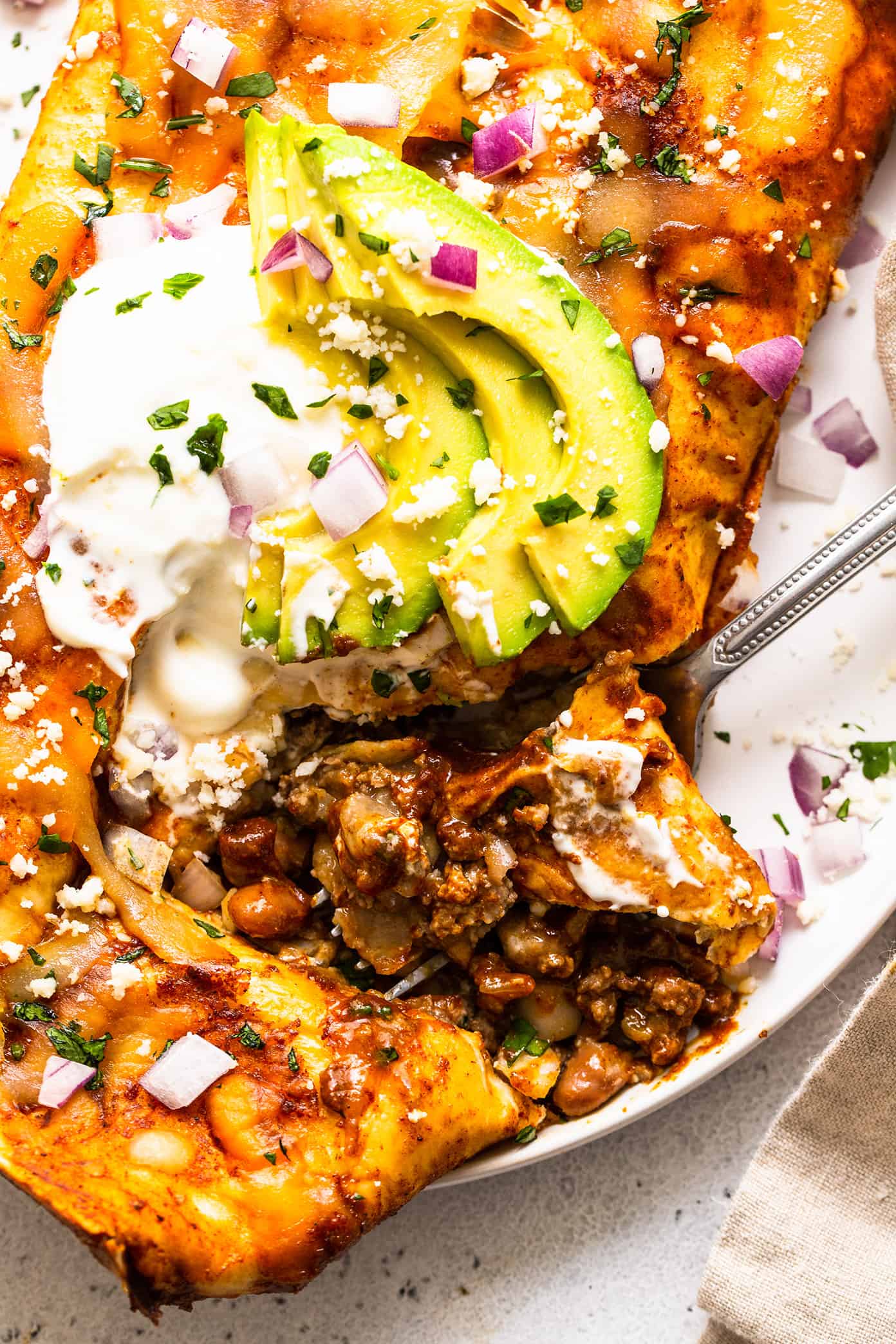 These delicious Tex-Mex-inspired enchiladas are loaded up with with tender ground beef (or plant-based meat), green chiles, onion, garlic, beans, cheese, corn or flour tortillas, and my favorite red enchilada sauce. When baked to warm and melty perfection, and served up with a few slices of avocado and perhaps a sprinkling of fresh cilantro and crumbled cotija cheese, they are always a crowd fave! Freezer and make ahead instructions included. | gimmesomeoven.com Beef enchilada closeup with avocado and sour cream