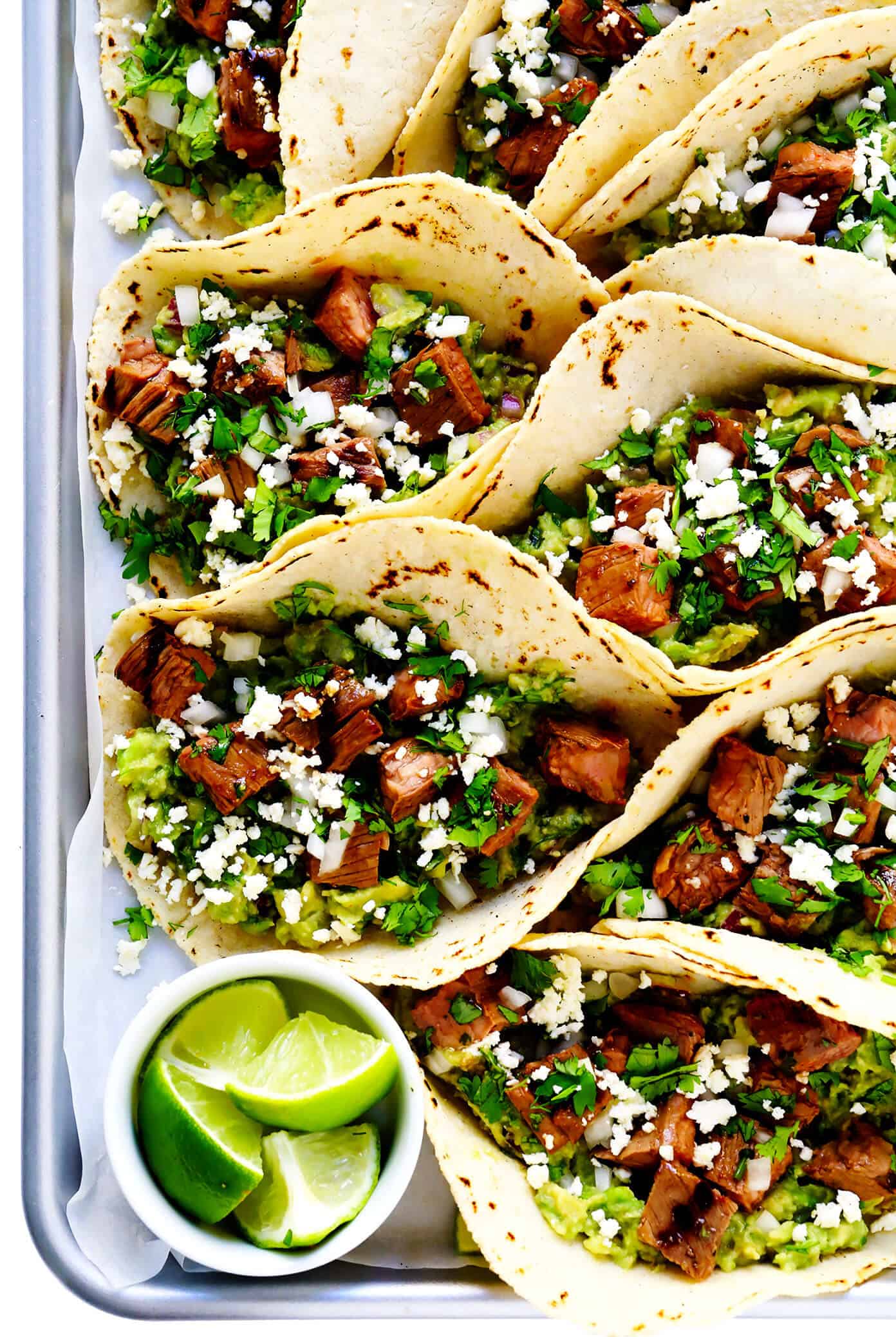 My favorite recipe for street-style Mexican carne asada tacos -- made with tender marinated grilled steak, soft corn tortillas, guacamole or sliced avocado, and whatever other toppings sound good! | gimmesomeoven.com #carneasada #beef #steak #tacos #mexican #avocado #guacamole #glutenfree #dinner Tray of Carne Asada Tacos with Lime, Guacamole and Queso Fresco
