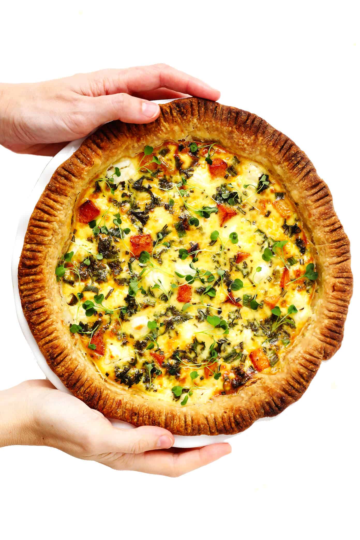 This easy-to-make butternut squash quiche is filled with garlicky kale, goat cheese, rosemary, creamy eggs, and baked in an all-butter pie crust. It's the perfect vegetarian recipe for weekend or holiday brunch and SO delicious. | gimmesomeoven.com #butternut #squash #quiche #breakfast #brunch #egg #bake #vegetarian #thanksgiving #christmas Holding a whole pan of Butternut Squash, Kale and Goat Cheese Quiche