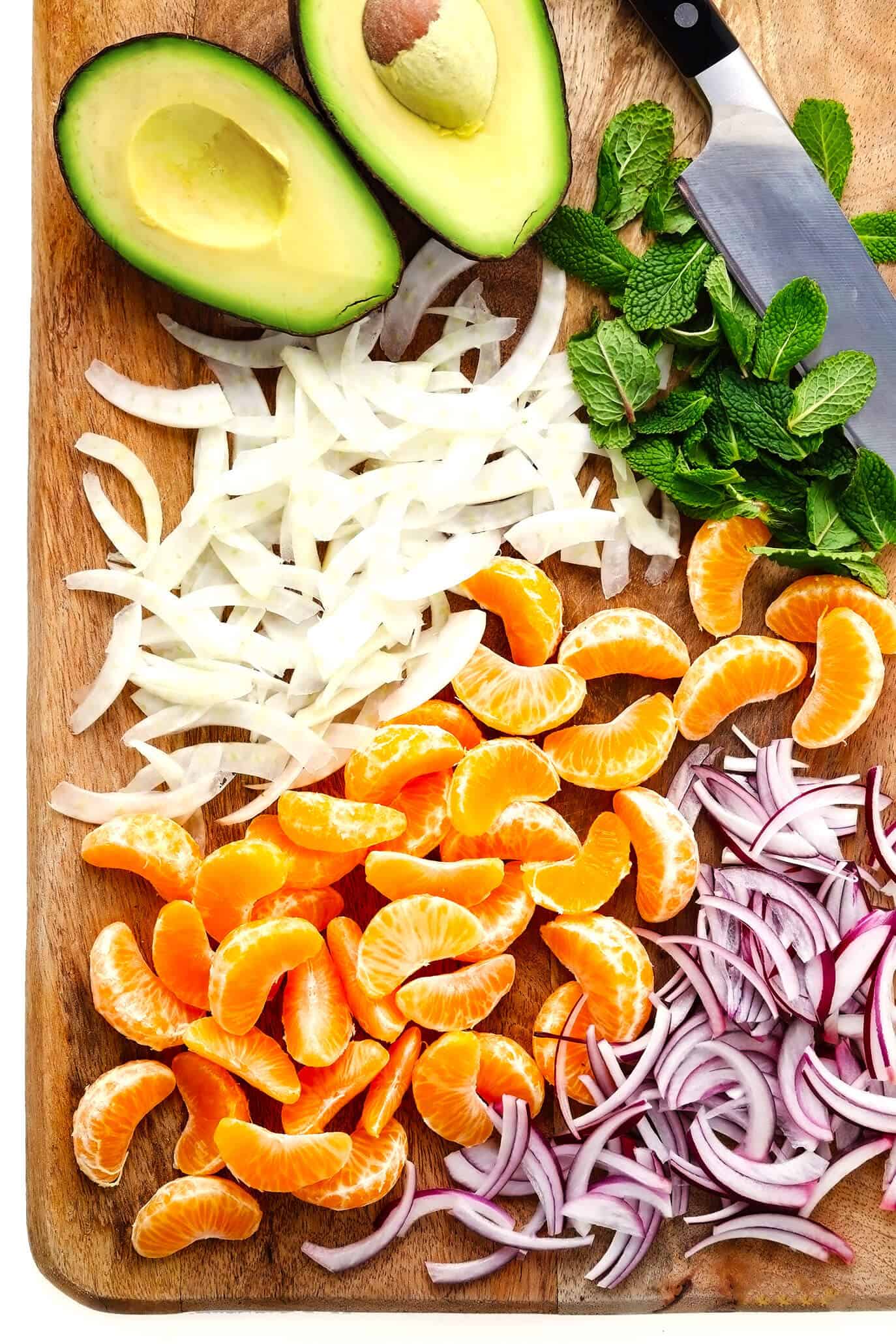 This Orange, Fennel and Avocado Salad recipe is quick and easy to make with fresh spring greens and mint, fresh clementines, toasted almonds, and a quick honey-citrus dressing. It's naturally vegan, vegetarian and gluten-free, but feel free to add in an extra protein (such as salmon, chicken or shrimp) if you'd like! | gimmesomeoven.com #salad #orange #citrus #avocado #healthy #glutenfree #vegan #vegetarian #dinner #winter Cutting board with fresh avocado, fennel, red onion, mint and clementine oranges