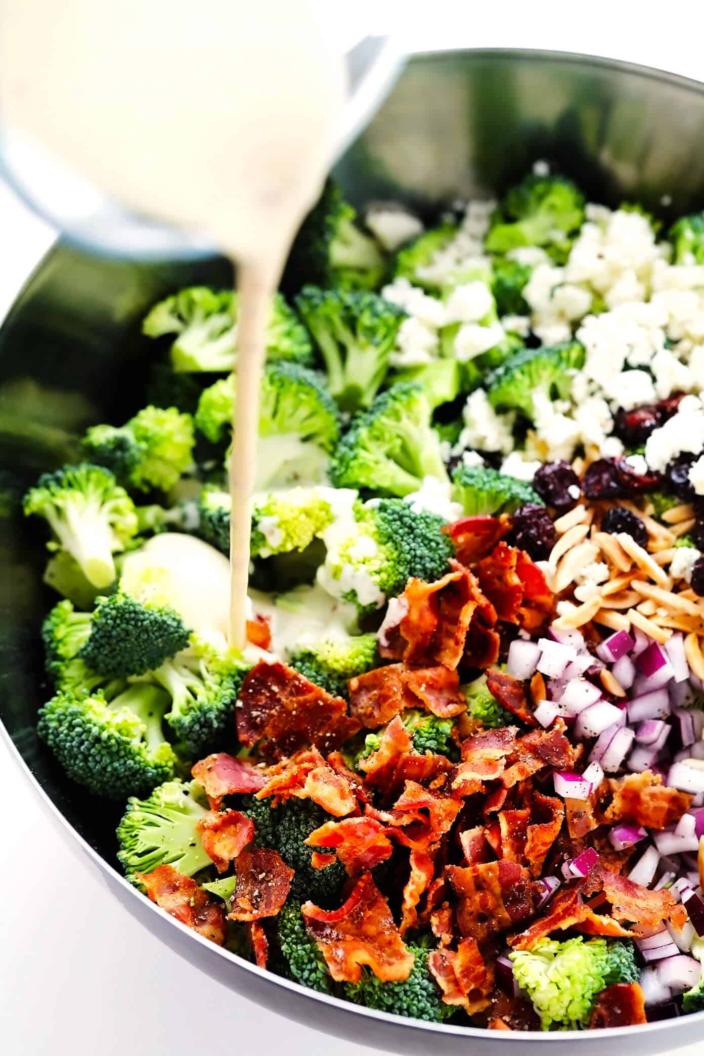 This classic broccoli salad recipe is made with fresh broccoli florets, crispy bacon, dried cranberries, toasted almonds, red onion, feta cheese, and a lightened-up creamy dressing. It's the perfect make-ahead side dish for picnics and potlucks and is always a crowd fave! | gimmesomeoven.com Pouring creamy dressing into broccoli salad