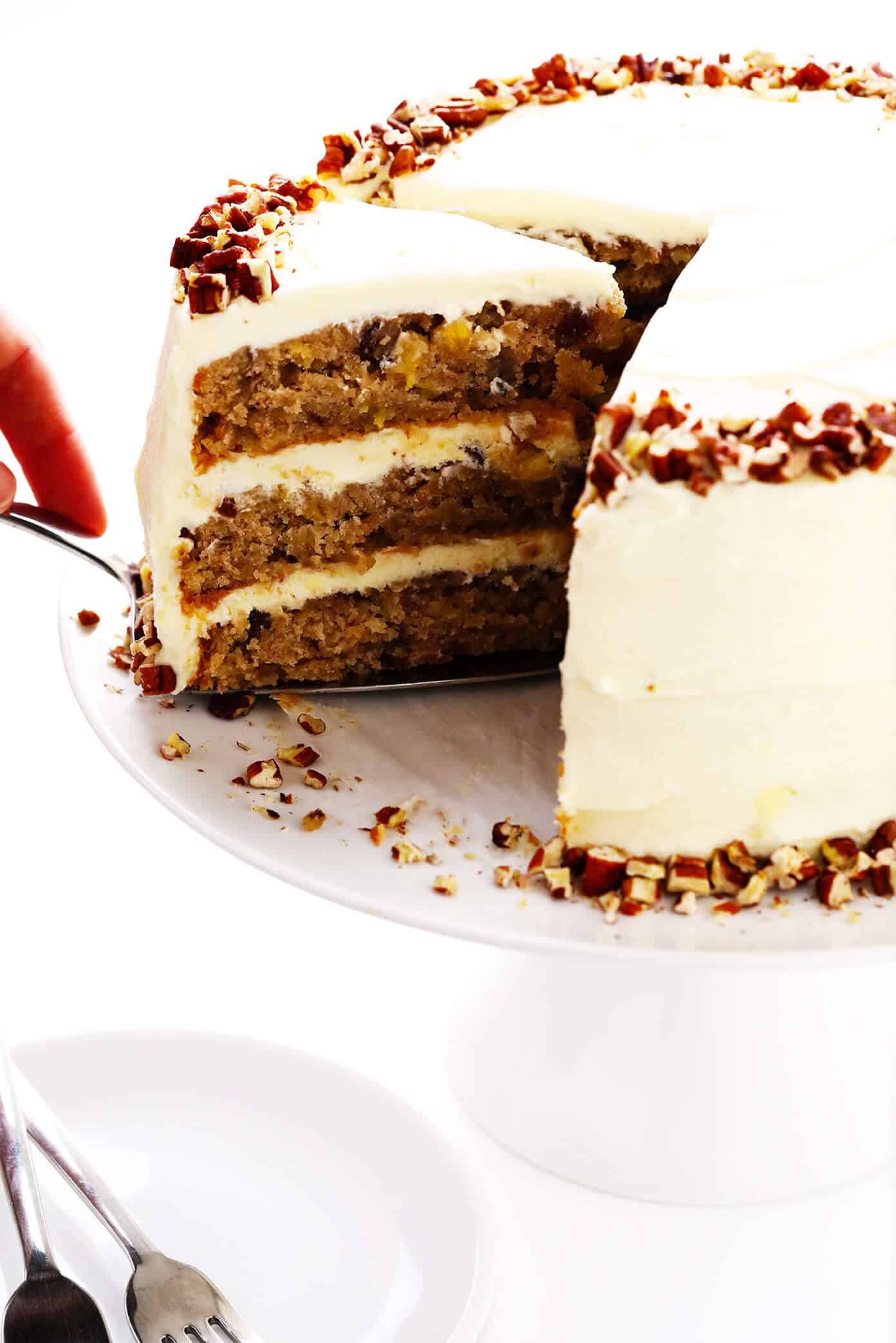 This delicious hummingbird cake recipe features ultra-moist pineapple banana cake layered with rich cream cheese frosting and a sprinkle of toasted pecans. It's the perfect dessert for spring, summer, or Easter and it's always a crowd fave! | gimmesomeoven.com #cake #dessert #hummingbird #jamaican #southern #creamcheese #frosting #icing #banana #pineapple #pecan Cutting a slice of hummingbird cake