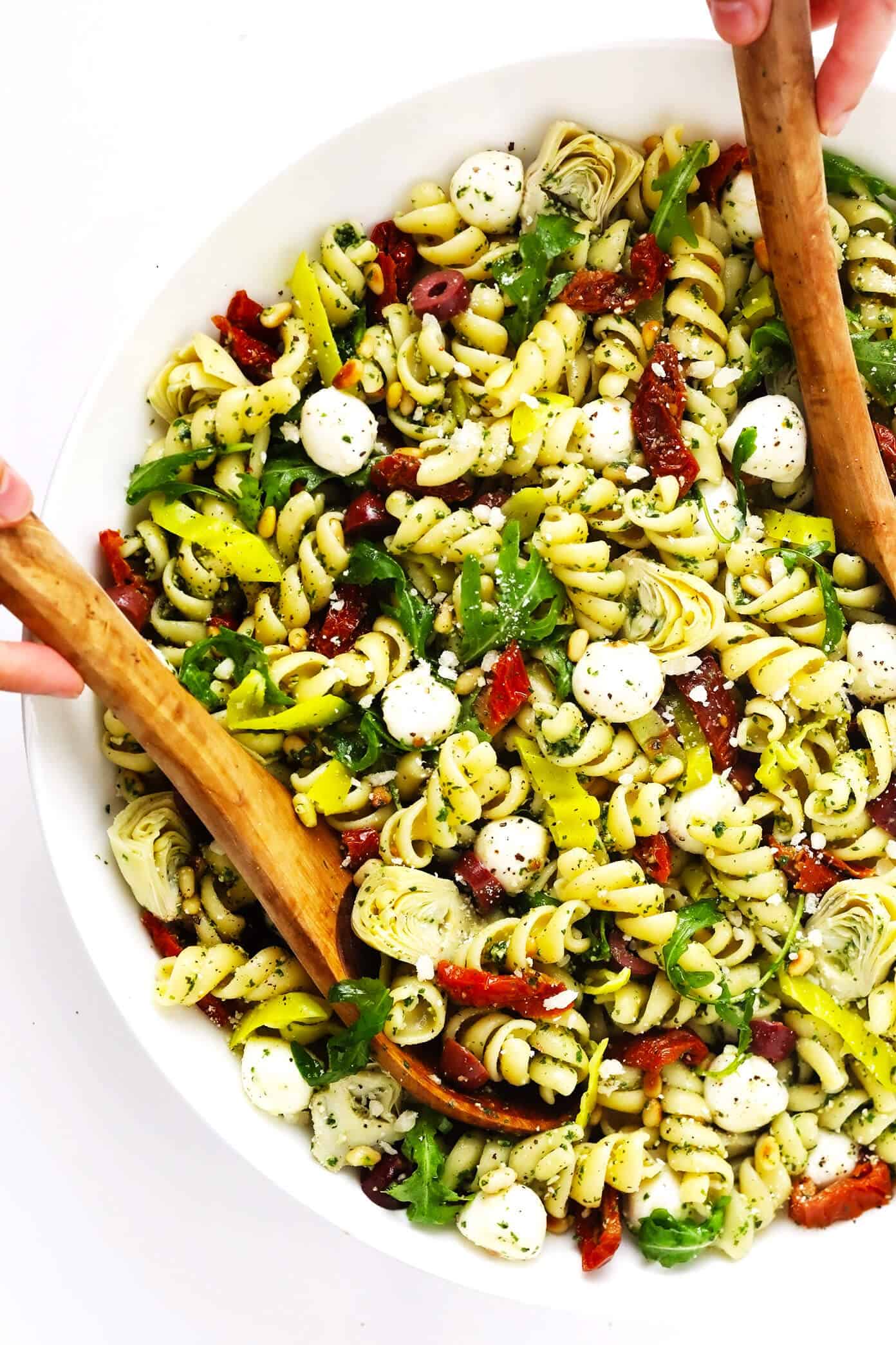 This Italian-inspired Pesto Pasta Salad recipe is loaded up with artichoke hearts, arugula, mozzarella, pepperoncini peppers, Kalamata olives, pine nuts and sprinkled with Parmesan. It's the perfect easy vegetarian side dish for summer grilling, parties and potlucks -- and easy to make gluten-free or vegan if you'd like too! | gimmesomeoven.com Italian Pesto Pasta Salad in Bowl with Tongs