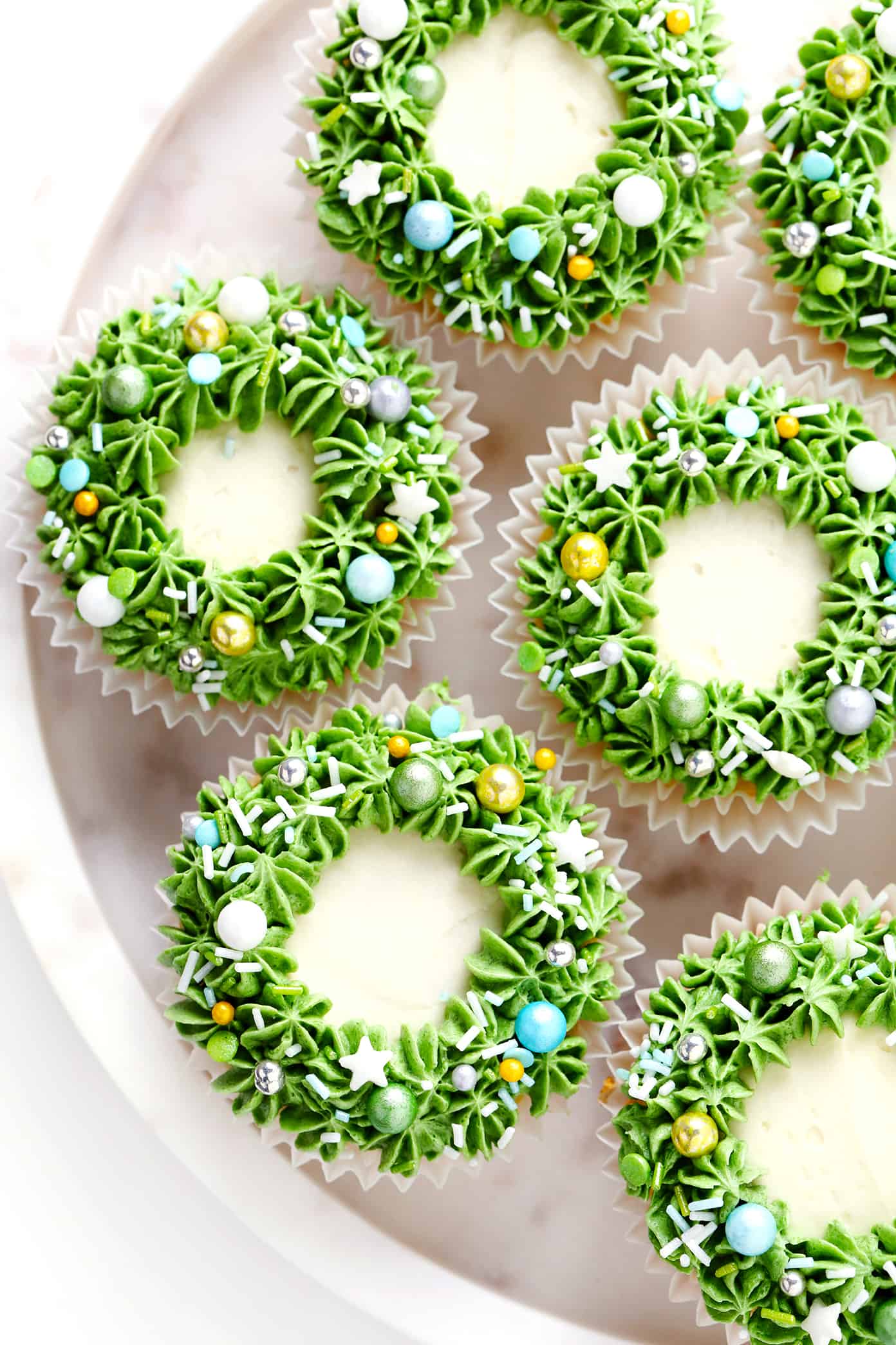 These cute Christmas cupcakes are made with soft and fluffy vanilla cupcakes, a simple vanilla buttercream frosting, and whatever fun sprinkles or toppings you choose. Decorating instructions and supply list included too for this fun holiday dessert! | gimmesomeoven.com Christmas Wreath Cupcakes with Sprinkles on Plate