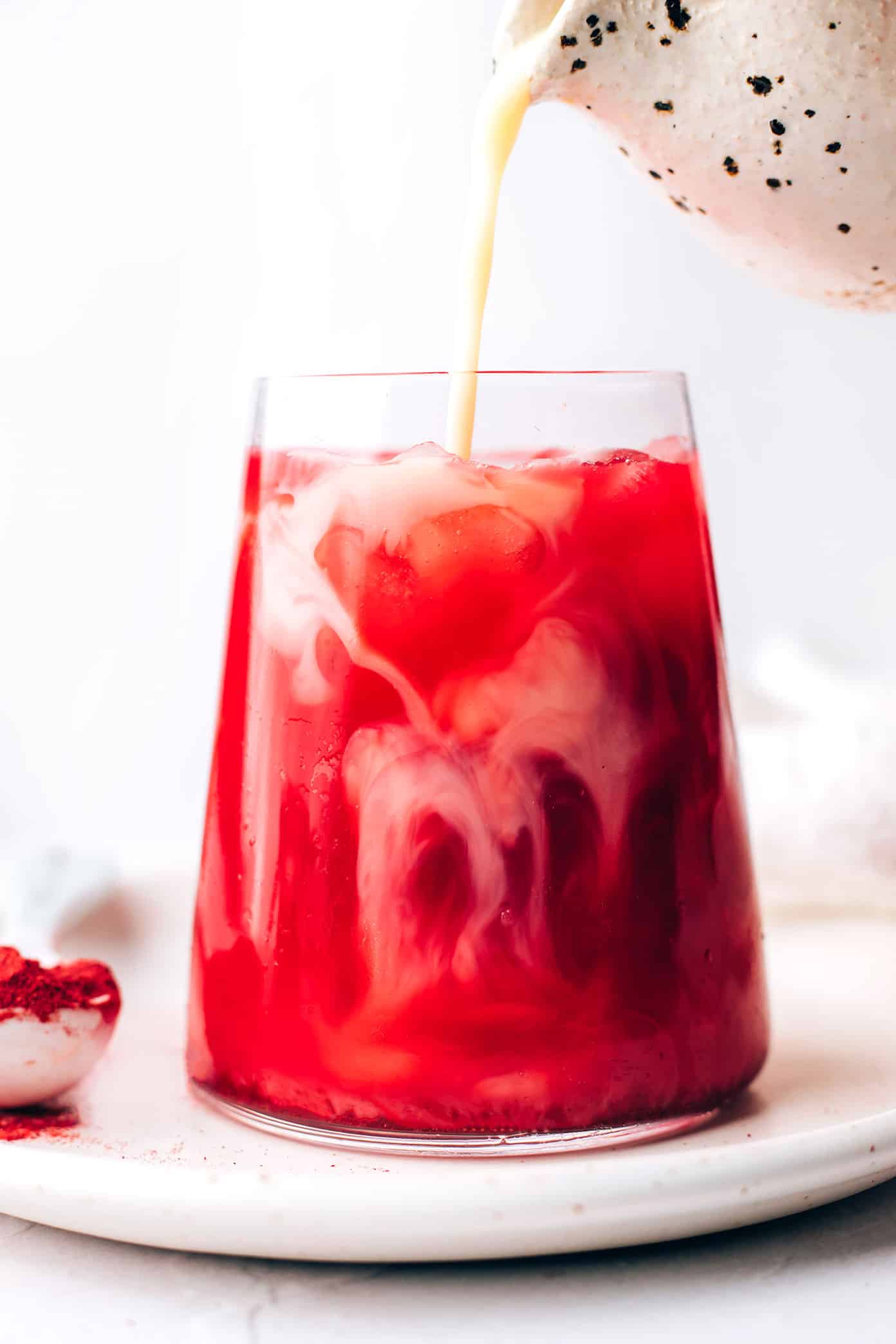 This nutrient-rich pink latte recipe is easy to make hot or iced and features a cozy mix of beetroot powder and warming spices (cardamom, cinnamon, ginger and cloves), your choice of milk and a hint of sweetener. Such cozy, vibrant and delicious drink! | gimmesomeoven.com Pouring milk into an iced beetroot latte