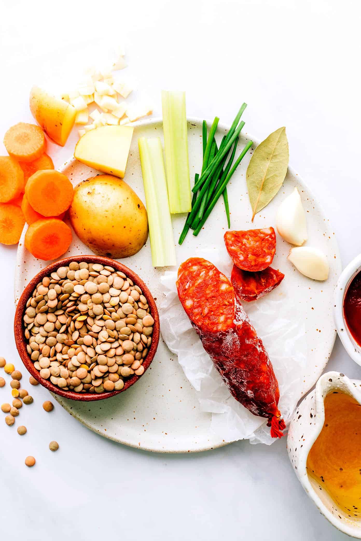 This Spanish Chorizo Lentil Stew recipe is incredibly easy to make, loaded up with veggies in a light paprika-tomato-garlic broth, and SO cozy and delicious. A great winter soup recipe for dinner when you're short on time! | gimmesomeoven.com Spanish Chorizo Lentil Soup ingredients on plate, including carrots, potato, garlic, celery, chives, bay leaf, garlic, tomato paste and broth